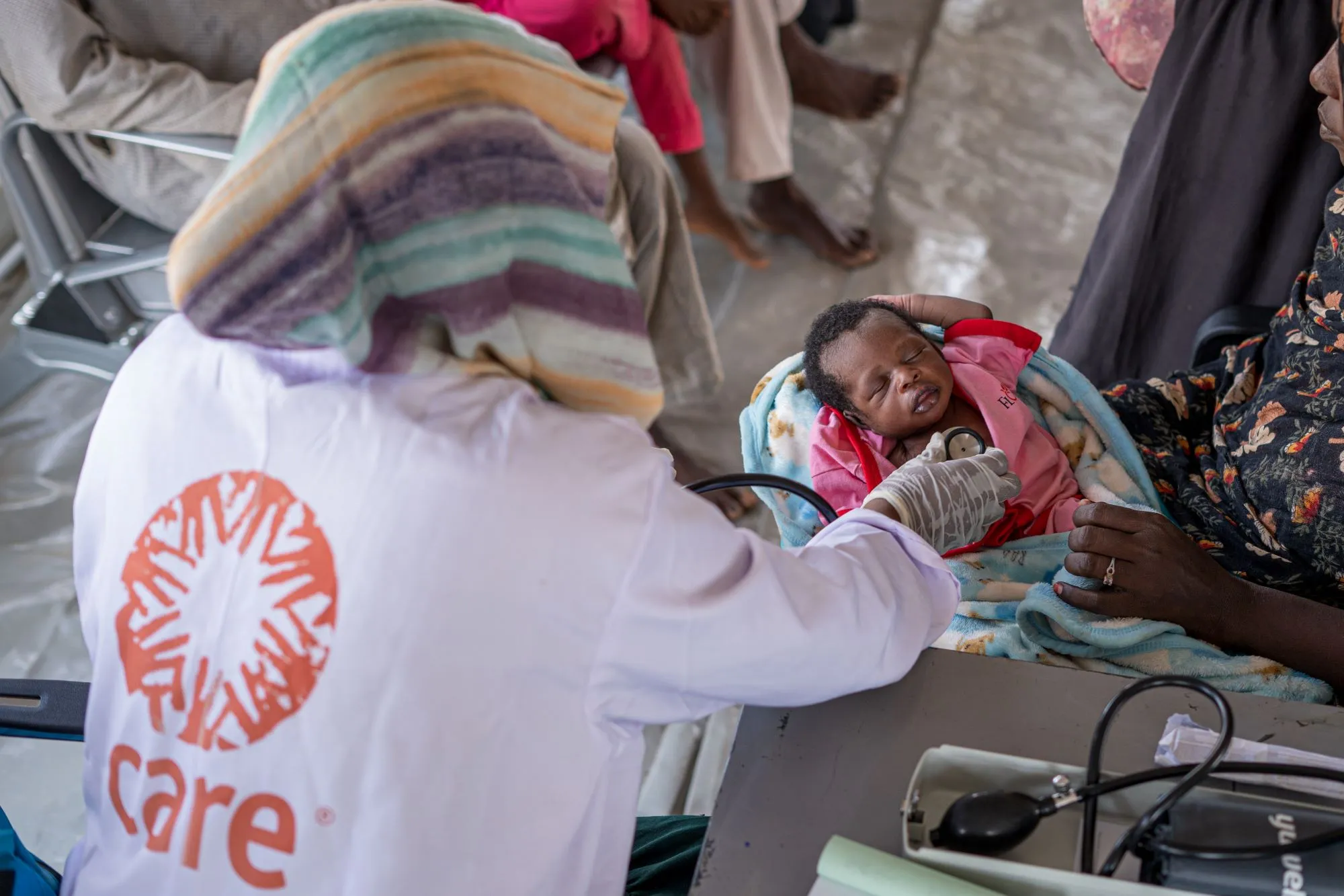 A healthcare worker wearing a CARE logo apron examines a child lying on a mother's lap.