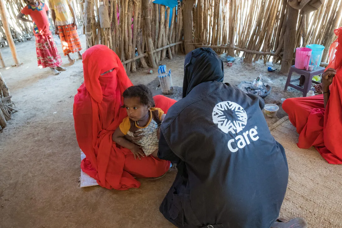 An aid worker wearing a dark jacket with the CARE logo kneels, examining a small child held by a woman fully veiled in red. This health check-up is taking place inside a rudimentary shelter made of wooden sticks in Sudan.