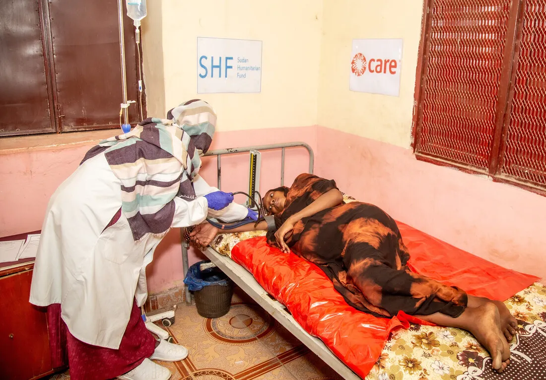 A female health worker, wearing a lab coat and headscarf, tends to a female patient lying on a bed with a red cover and receiving an IV drip. The wall displays signs for the SHF (Sudan Humanitarian Fund) and CARE, indicating vital medical support at this facility.