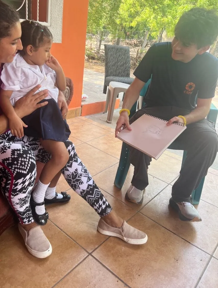 A young man in a CARE t-shirt shows a drawing in a sketchbook to a woman and a young child sitting on a porch in Honduras.