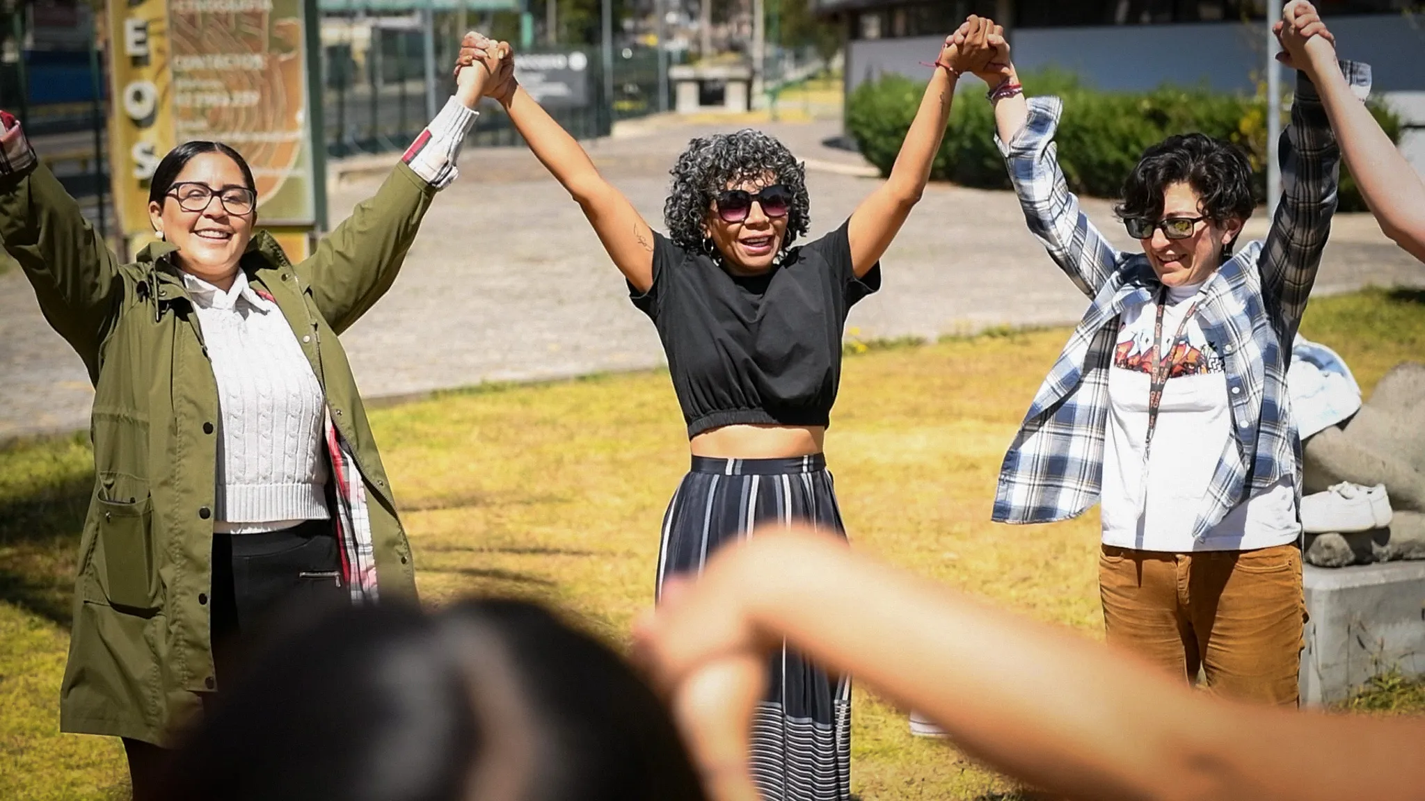 Three women stand outdoors holding hands and raising their arms together, smiling in a moment of unity during a community gathering.