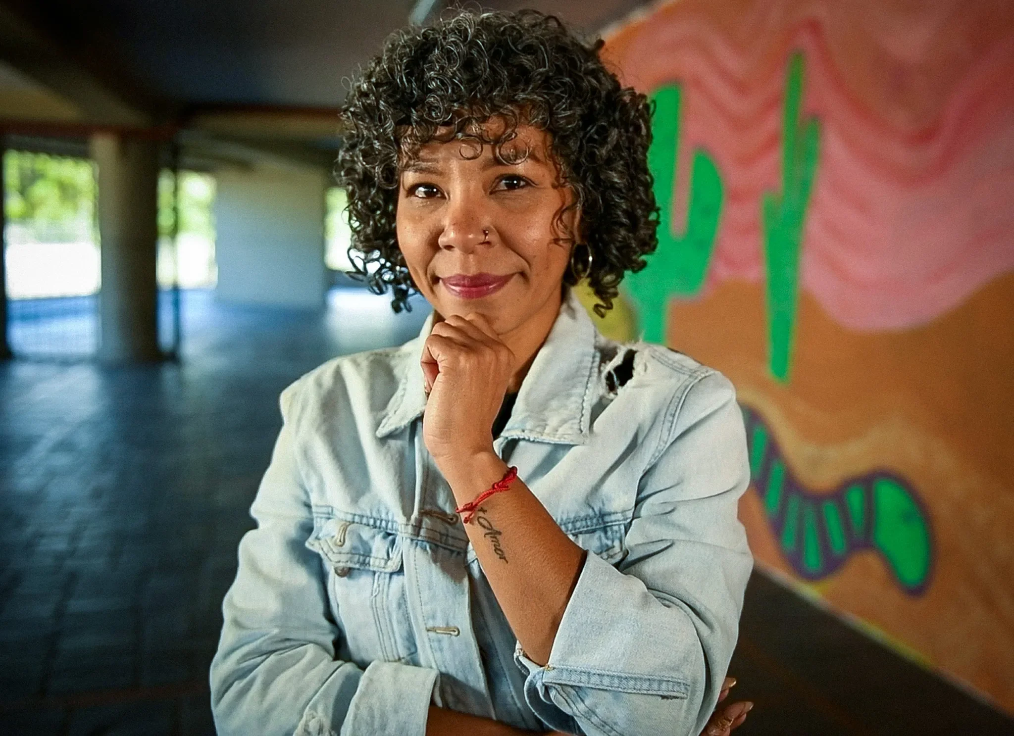 A woman with short curly hair stands resting her chin on her hand. Behind her are colorful murals.
