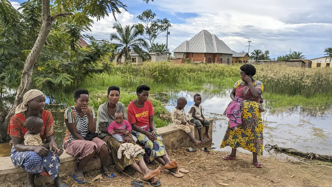 People seated outside by a lake. Palm trees and a small house can be seen in the background.