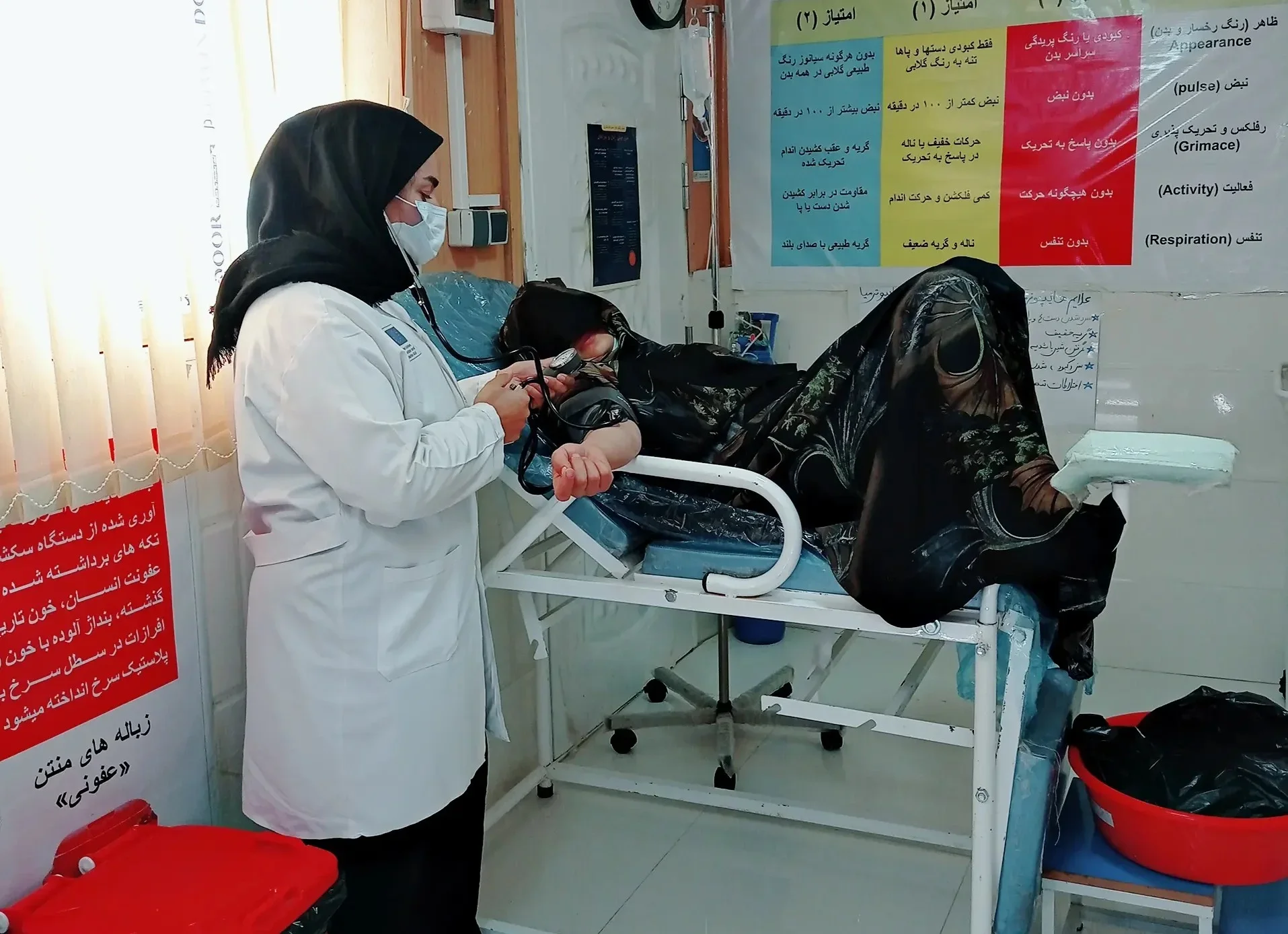 A midwife in a white coat and face mask checks the blood pressure of a pregnant patient lying on a medical bed in a clinic in Afghanistan.