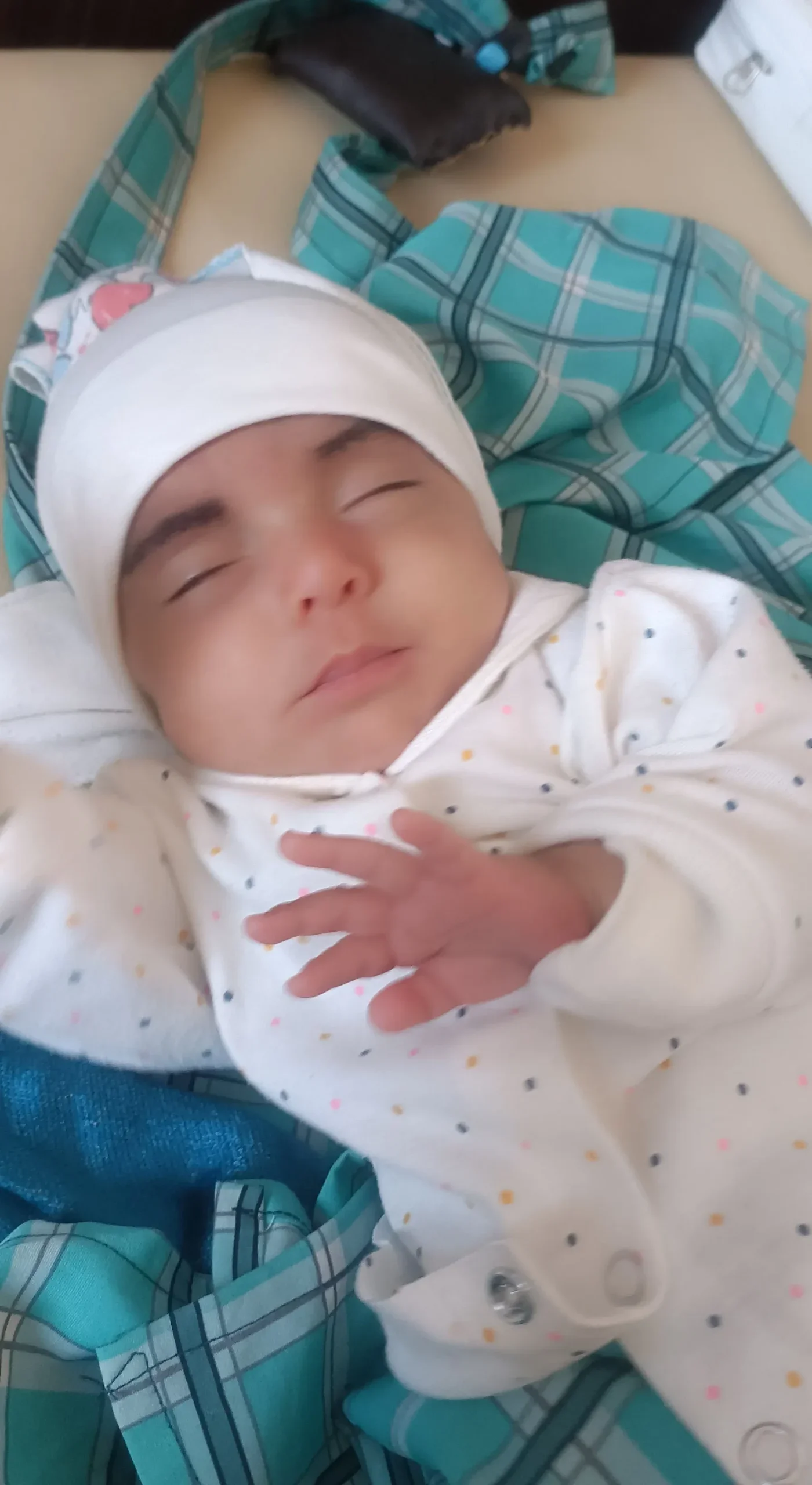 A close-up of baby Forozan, a newborn wrapped in a white polka-dot onesie and a white beanie, resting peacefully against a teal checkered blanket.