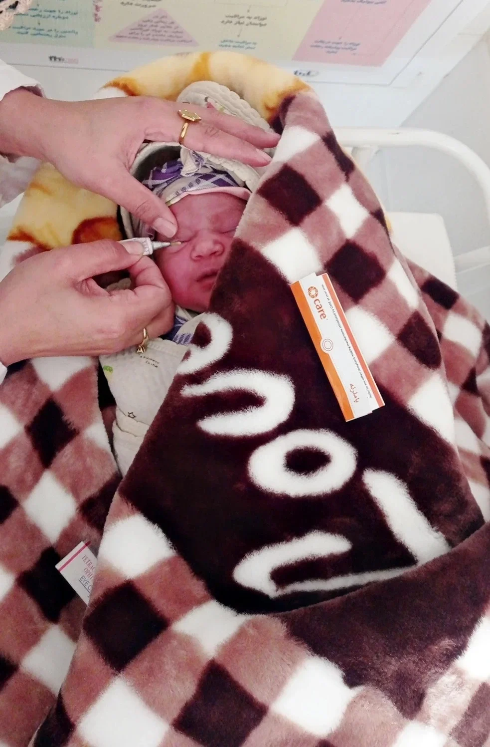 A healthcare worker's hands carefully apply eye drops to a newborn baby who is wrapped in a thick, brown and white checkered blanket at a CARE-supported clinic.