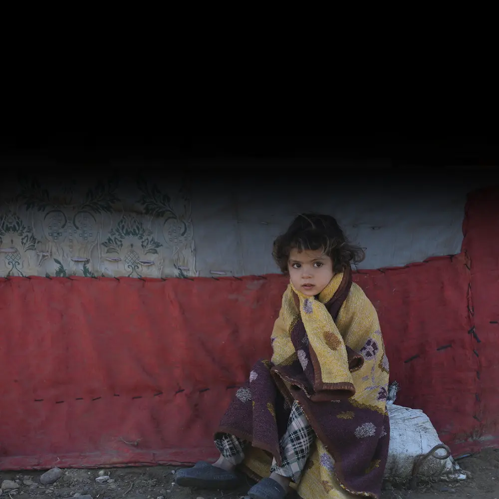 A young Syrian child huddles outside a tent in a fleece blanket.