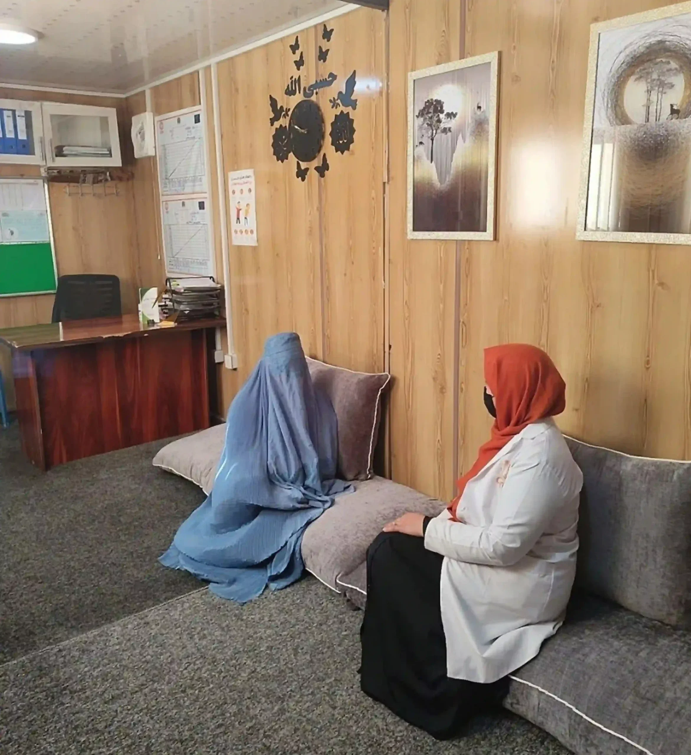 A female healthcare provider in a white coat and orange headscarf sits on a cushioned bench, engaged in a private counseling session with a patient wearing a blue burqa in a wood-paneled office in Afghanistan.