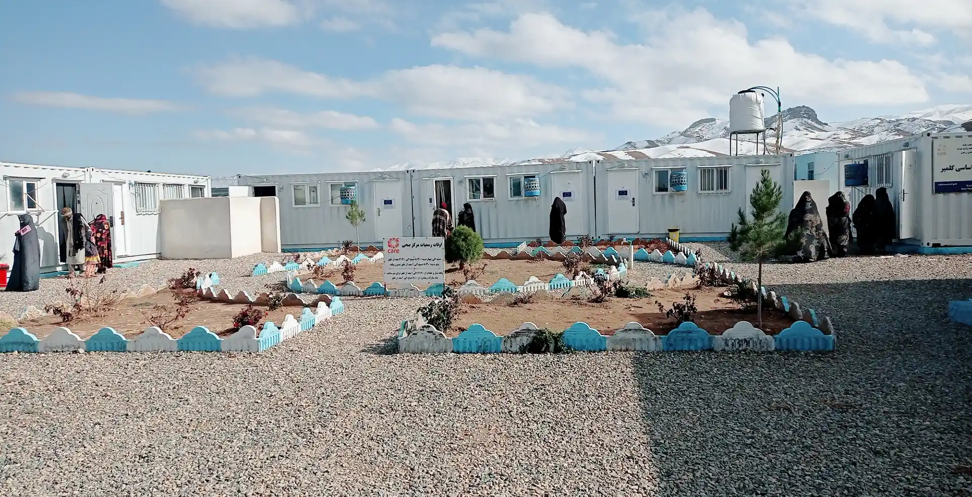 An outdoor view of the Kolmir Basic Health Center, a facility made of white shipping containers in a remote, rocky landscape with snow-capped mountains in the background. Patients walk near the entrance and small garden plots, while a prominent signboard featuring the CARE logo stands in the foreground.