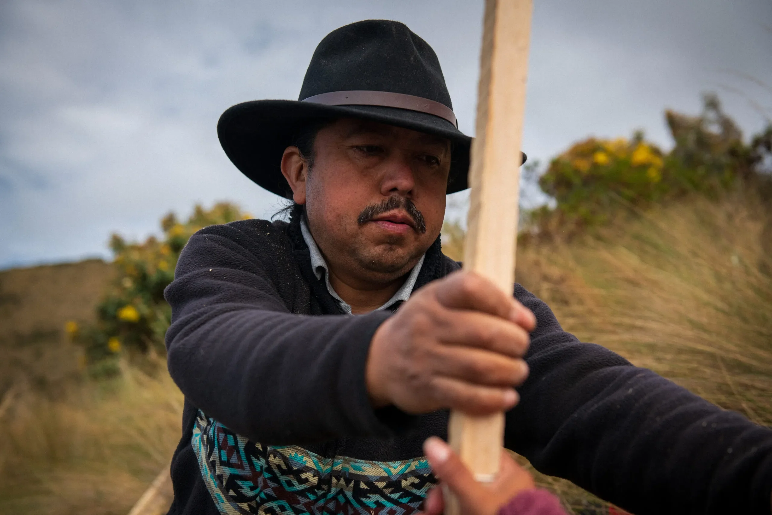 Close-up of Hilario Morocho cutting wooden stakes in the páramo to mark the placement of firebreaks, with another community member just out of frame.