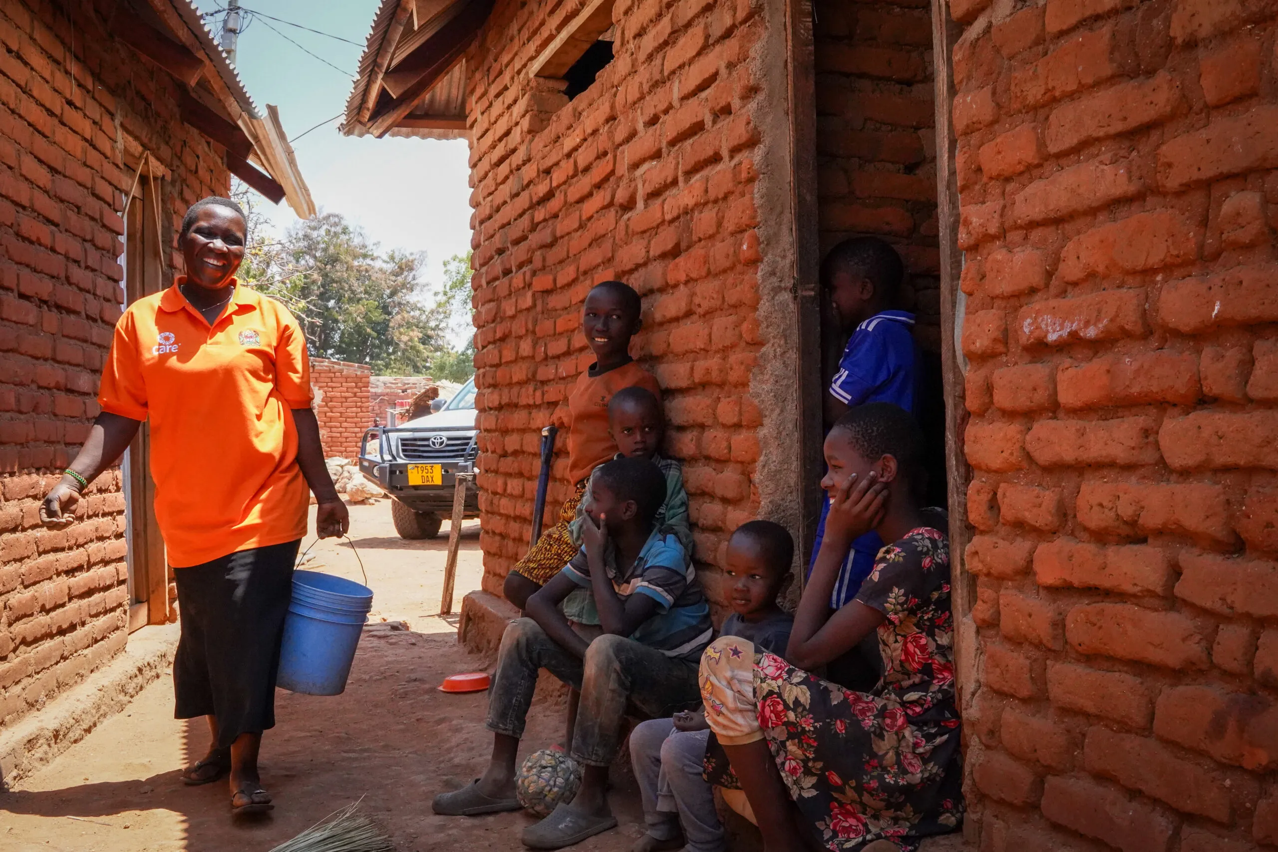 Sikujua carries a bucket between brick buildings in a Tanzanian village while children smile and watch her work.