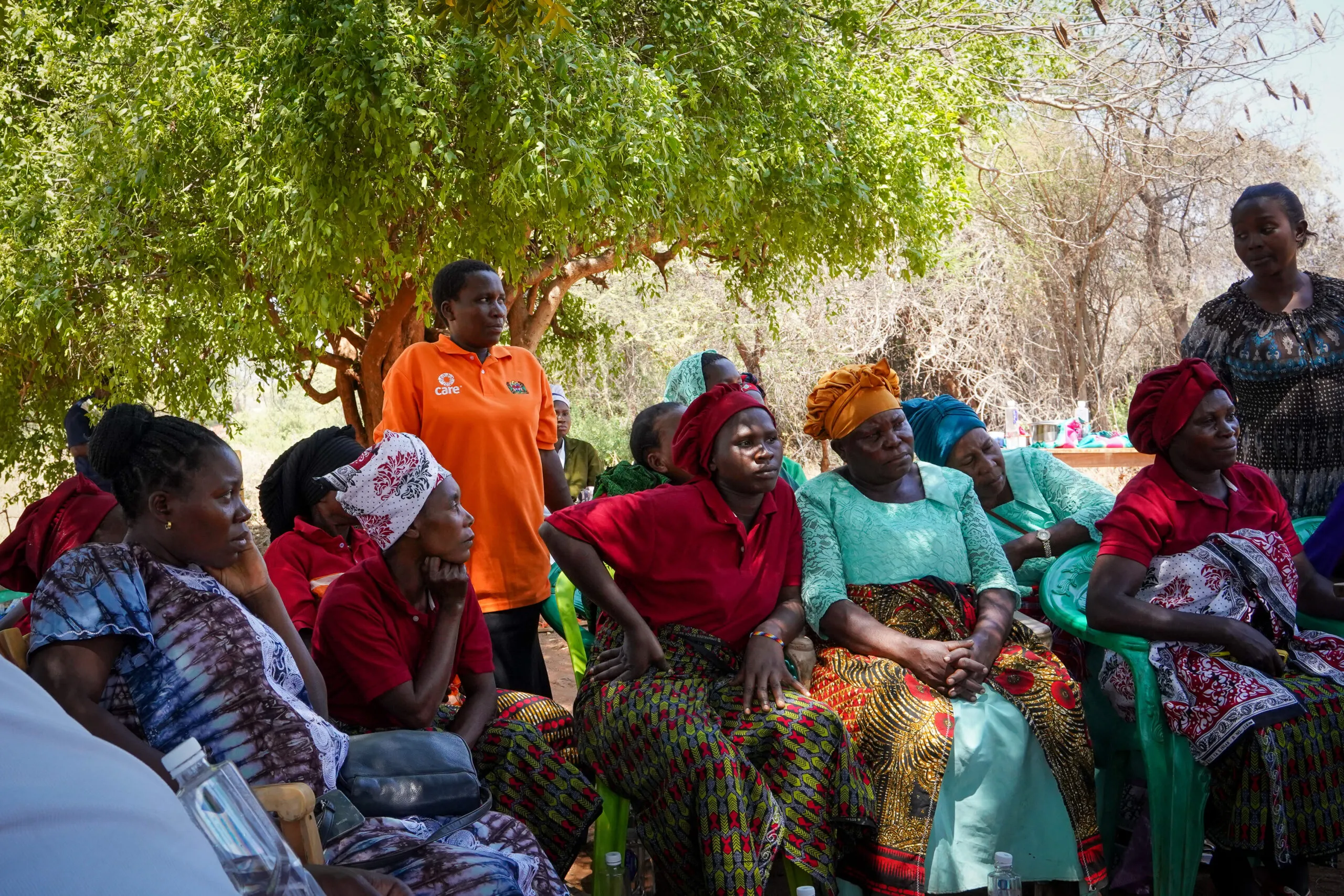 Women wearing colorful wraps and headscarves sit under a tree during a Village Savings and Loan Association meeting in Tanzania, with Sikujua Uhaula wearing a CARE shirt among them.