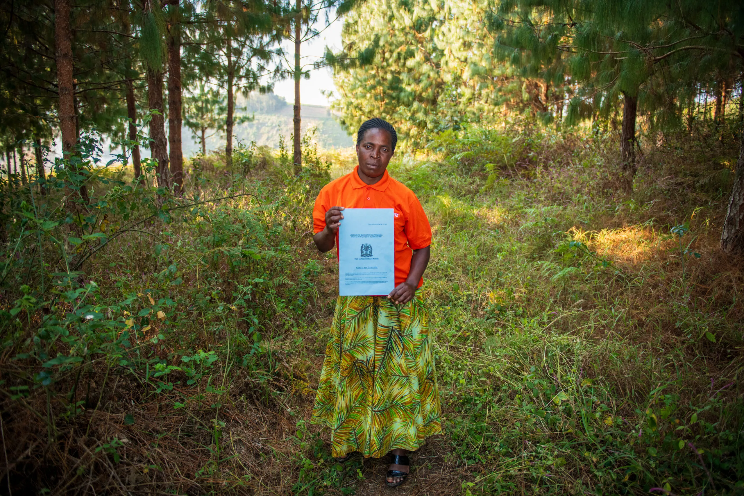 Yuditha Alimoti Sanga stands on her timber farm in Tanzania holding an official land ownership certificate while wearing a CARE shirt.
