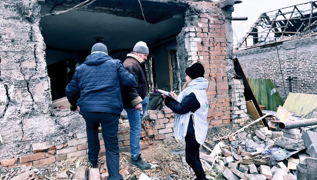 Three people in winter clothing inspect a partially collapsed brick building. Debris surrounds them in a scene of recent destruction.