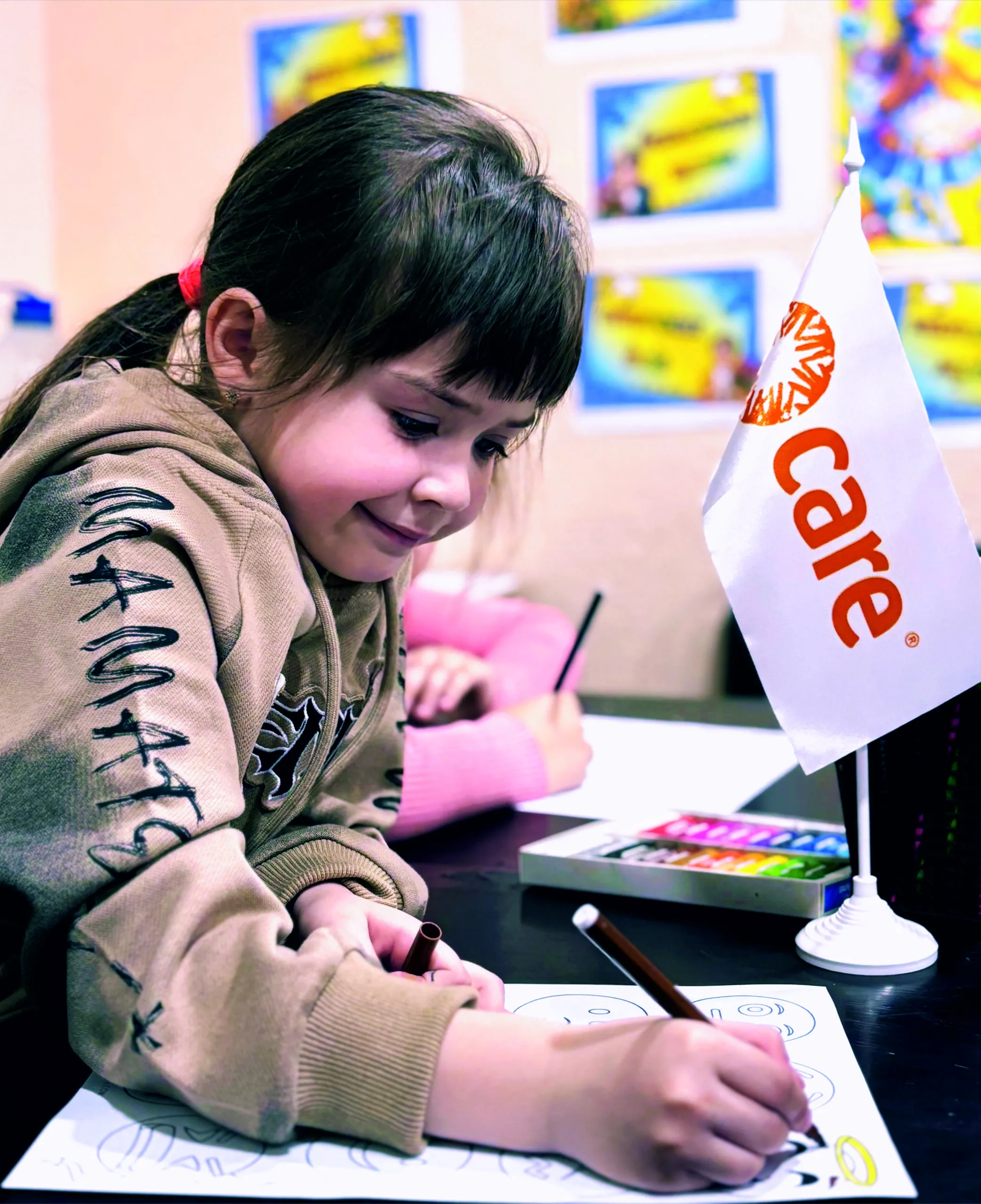 A young girl in a brown sweater is smiling and drawing at a table. A small flag reads 