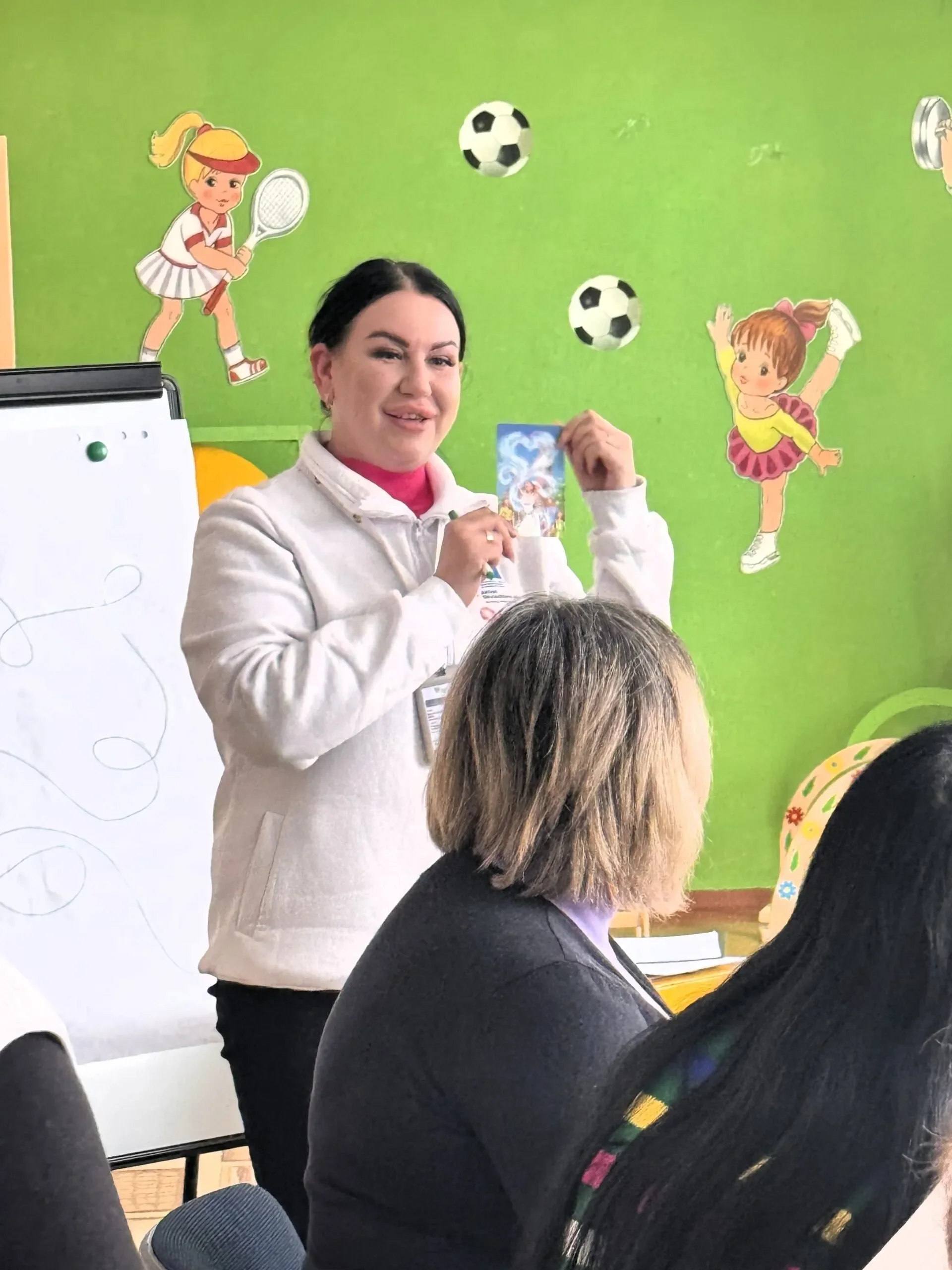 A woman holds a card while presenting to a group in a brightly decorated room with sports-themed wall art. She smiles, creating an upbeat atmosphere.