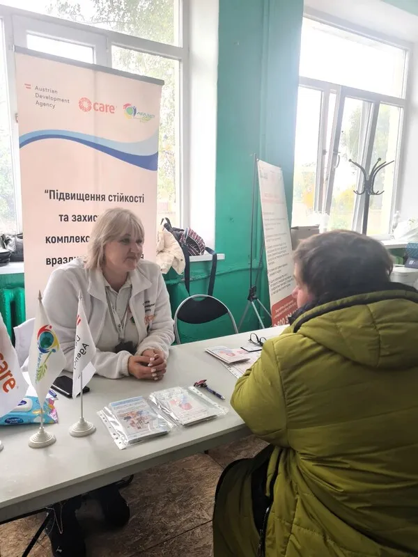 A woman in a white coat sits at a table with brochures, talking to a person in a green jacket. Banners behind them share information on support resources.
