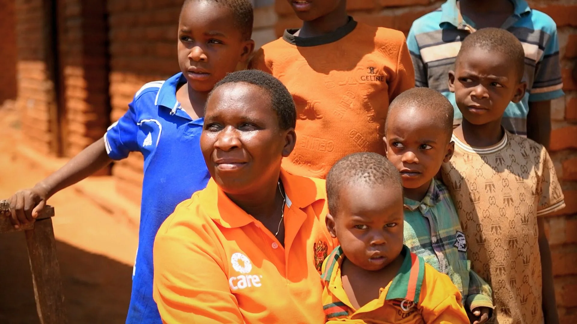 Sikujua Uhaula sits with her family in Tanzania’s Iringa Region, wearing an orange CARE shirt, holding a young child and looking into the distance.