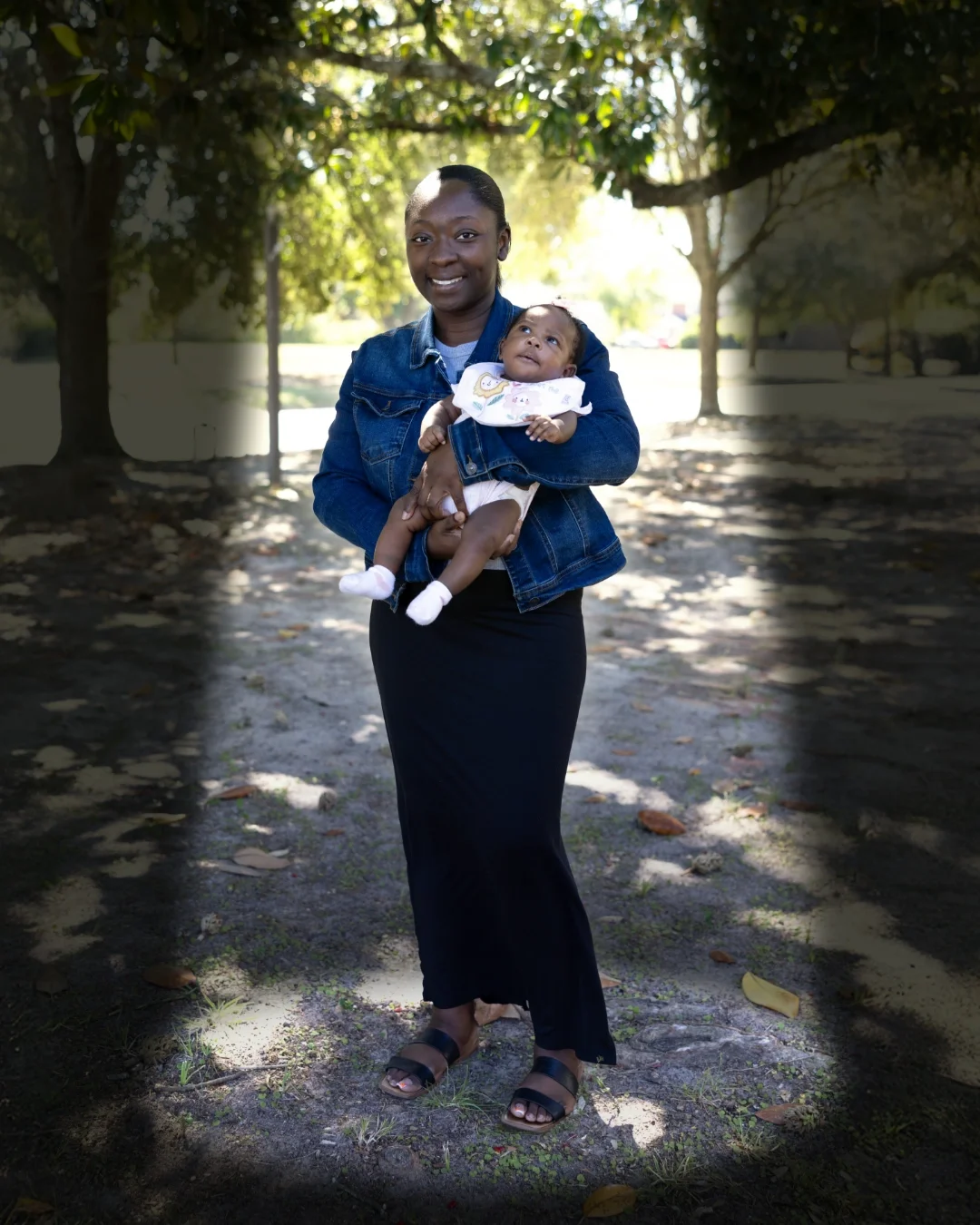 A Black American woman standing under a tree smiles while holding her young baby.
