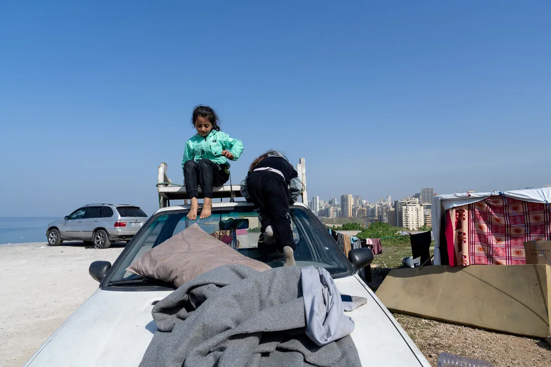 Two children sit on a car roof surrounded by makeshift textiles, with a city skyline in the background under a clear blue sky, evoking resilience.