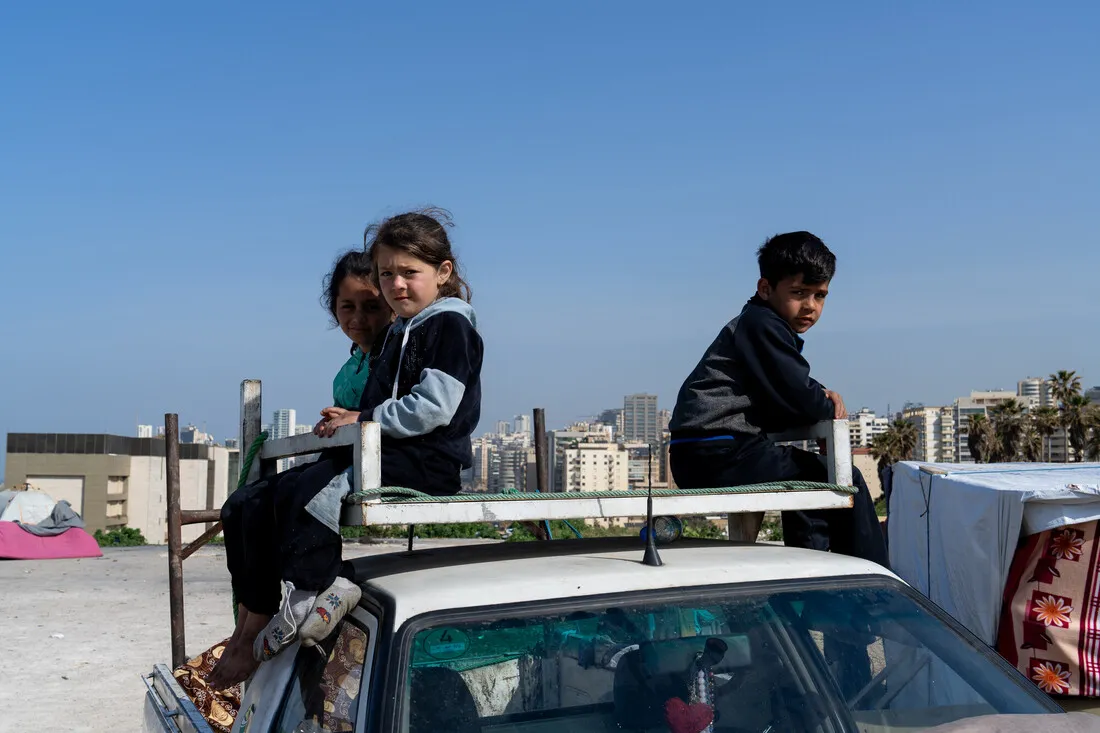Three children sit on a rooftop car rack in an urban area. The sky is clear, and city buildings form the backdrop. Their expressions are serious.