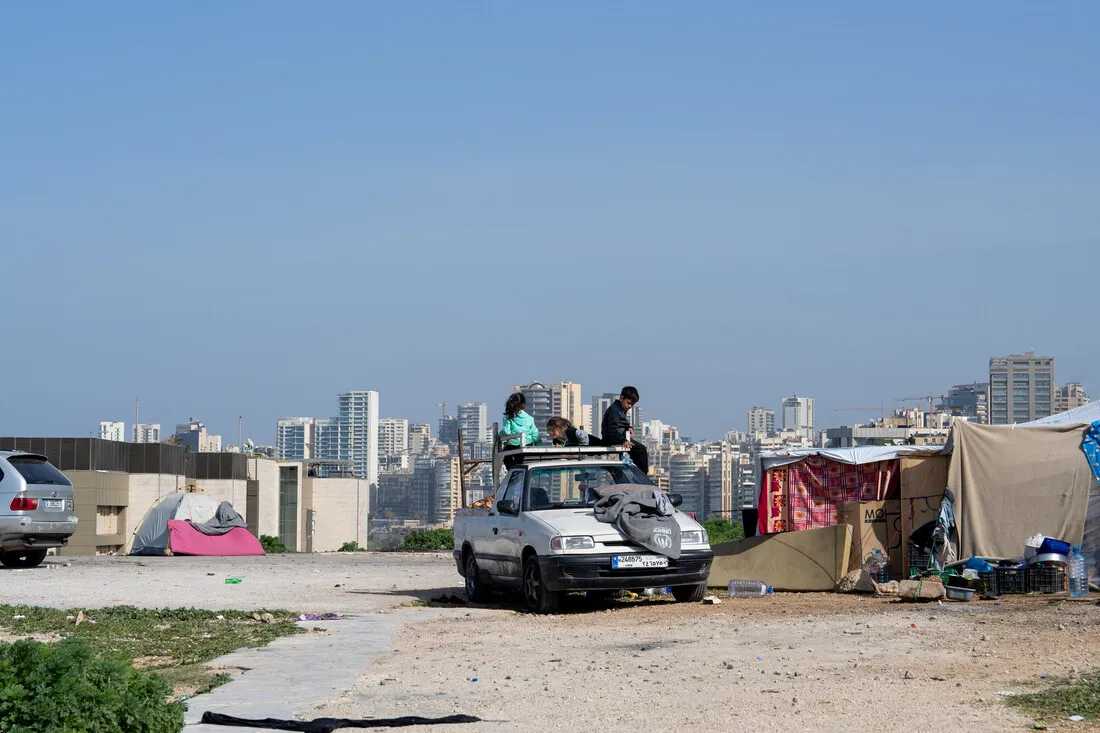 A car with people on top parked near makeshift tents on a dirt area. In the background, a cityscape with tall buildings under a clear blue sky.