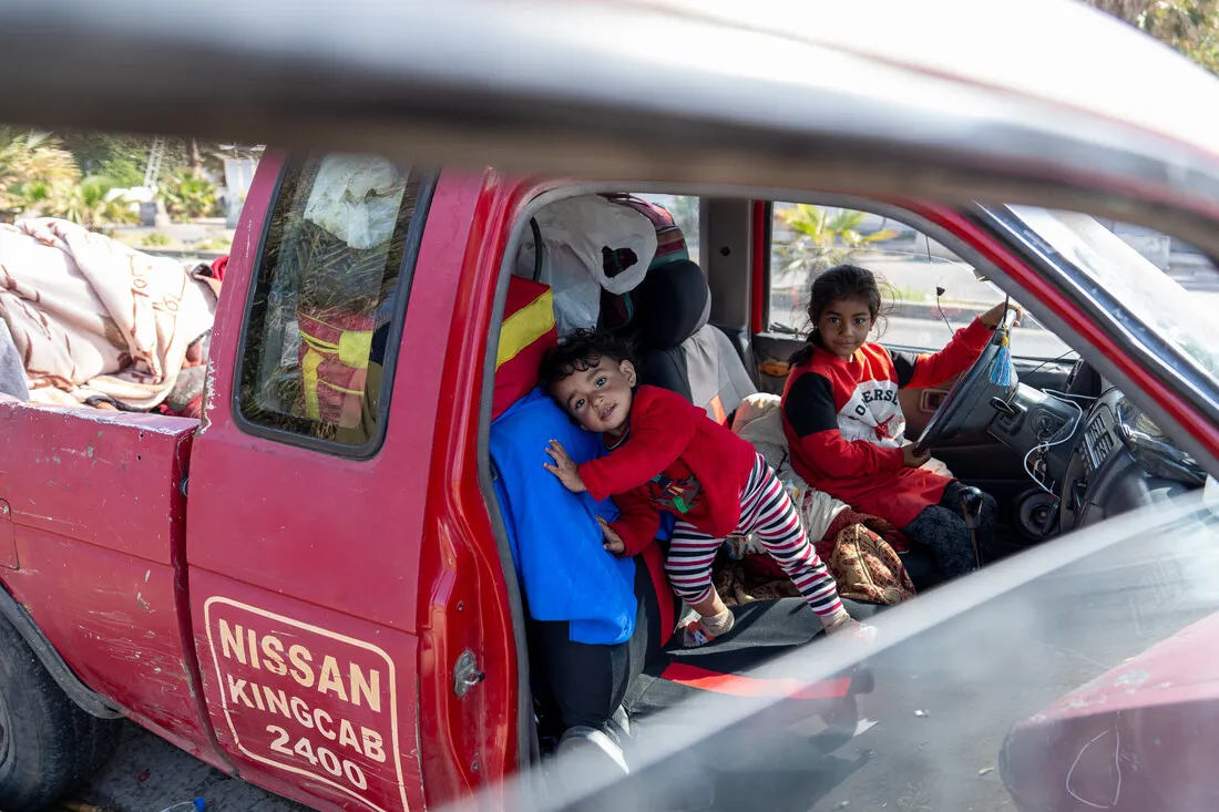 Two young children sit inside an old, red Nissan truck filled with belongings. One child leans playfully on the seat, while the other sits in the driver's seat.