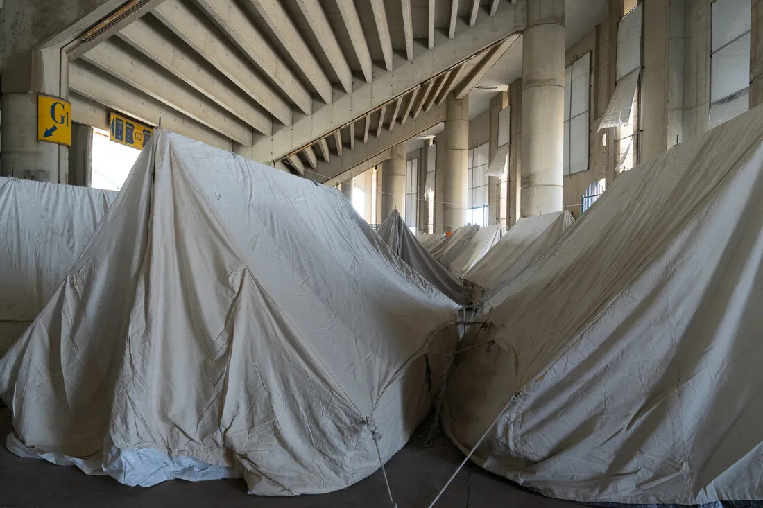 Rows of beige tents are set up under a large concrete structure with exposed beams and pillars, creating a temporary shelter space. The atmosphere is utilitarian.