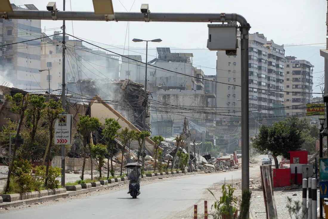 A motorcycle travels down a deserted street lined with debris and buildings damaged by conflict. Smoke rises from the ruins, conveying destruction and desolation.