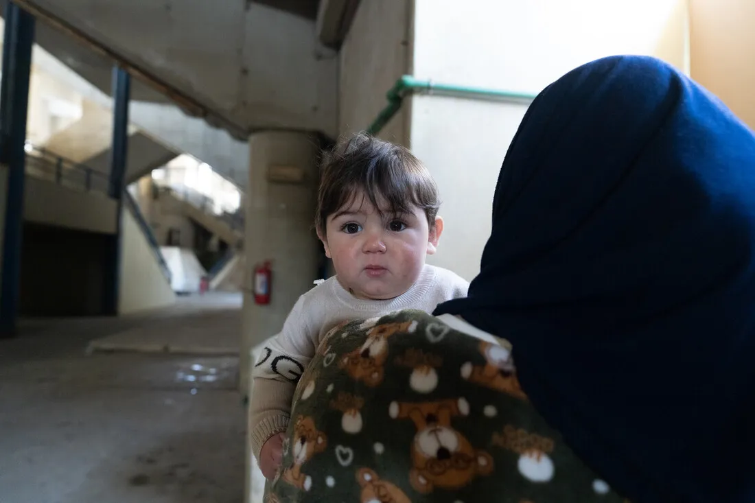 A woman in a blue headscarf carries a baby in a teddy bear blanket. They are in a dimly lit concrete structure; the baby gazes calmly at the camera.