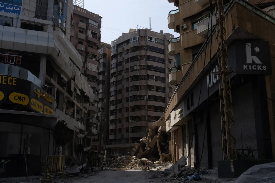 Urban street with tall, damaged buildings and debris blocking the road. The scene conveys a sense of desolation and abandonment in a city setting.