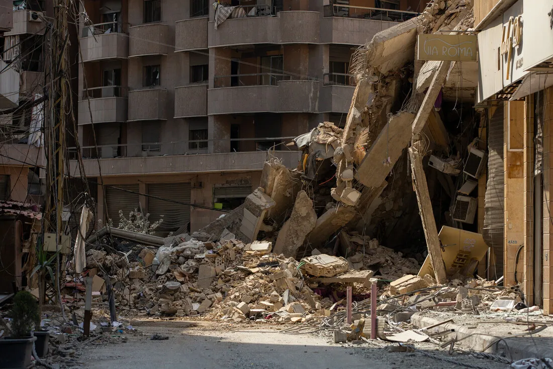 Collapsed building with debris and rubble in an urban street. Nearby structures show signs of damage. The scene conveys destruction and chaos.