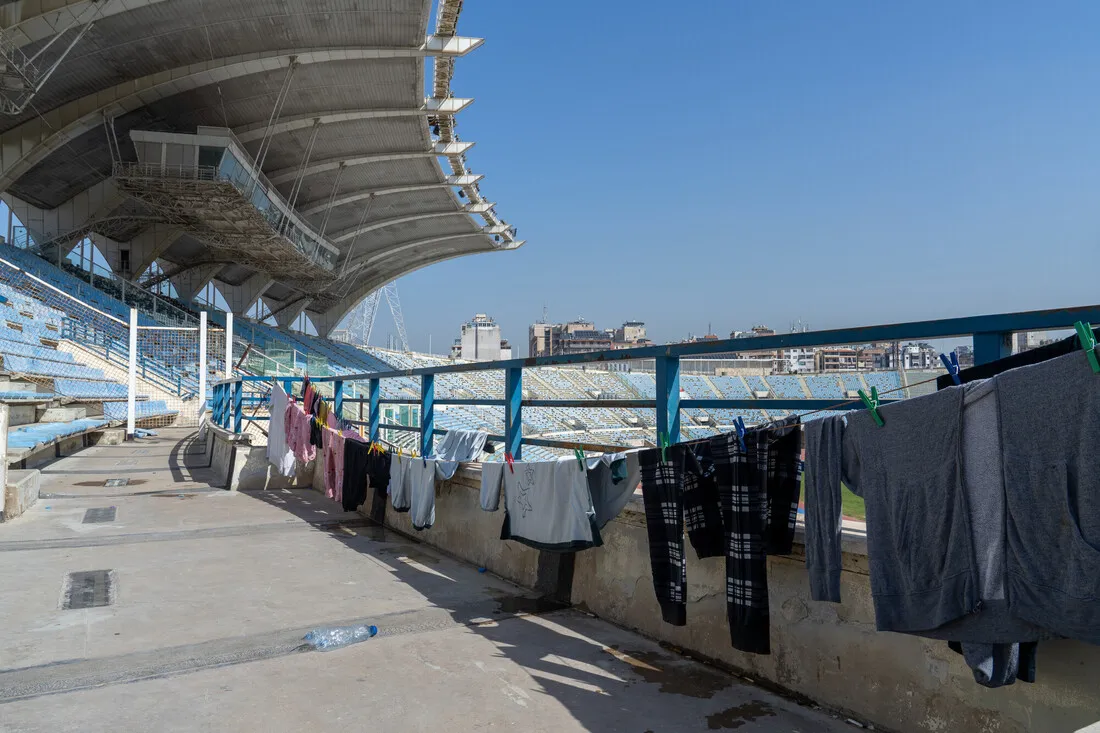 Clothes hang on a railing under a large stadium roof, with empty blue seats in the background and a cityscape under a clear blue sky.