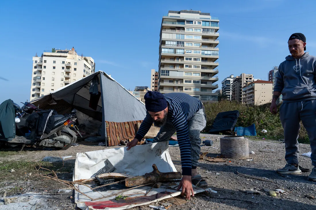 Two people in casual clothing work near a makeshift tent with scattered belongings in an urban area, with high-rise buildings in the background. The scene conveys resilience and resourcefulness.