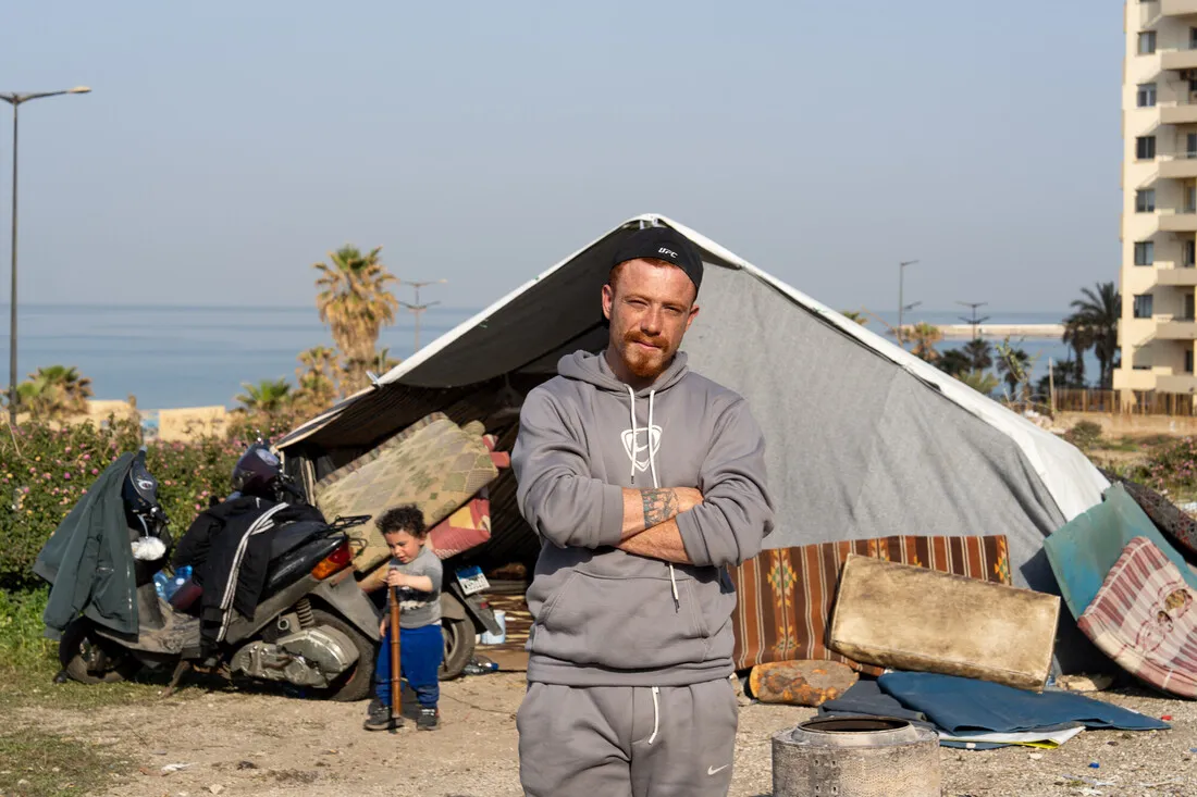 A man stands with arms crossed in front of a makeshift tent by the seaside, with a child and a parked scooter nearby. The mood is resilient.
