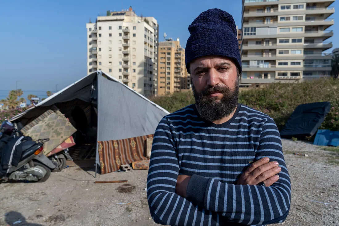 Man in beanie and striped sweater stands with crossed arms in front of a makeshift tent. Urban buildings are visible in the background, under clear skies.