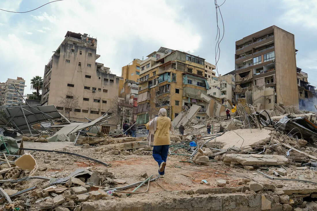 A person in a hooded jacket walks through rubble and debris of a collapsed building in an urban area. Nearby damaged buildings stand under a cloudy sky. The scene conveys devastation and resilience.