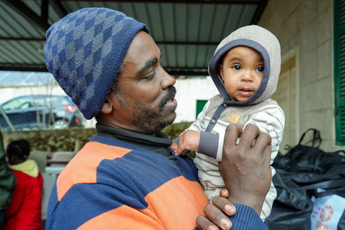 A man in a blue beanie and striped sweater holds a baby in a gray hoodie, looking at each other affectionately. Background has outdoor items.