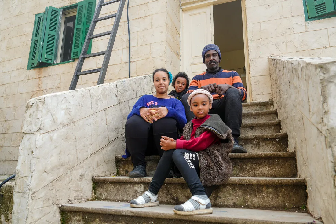 A family of four sits on stone steps outside a building with green shutters. They appear peaceful and close, creating a warm, familial atmosphere.