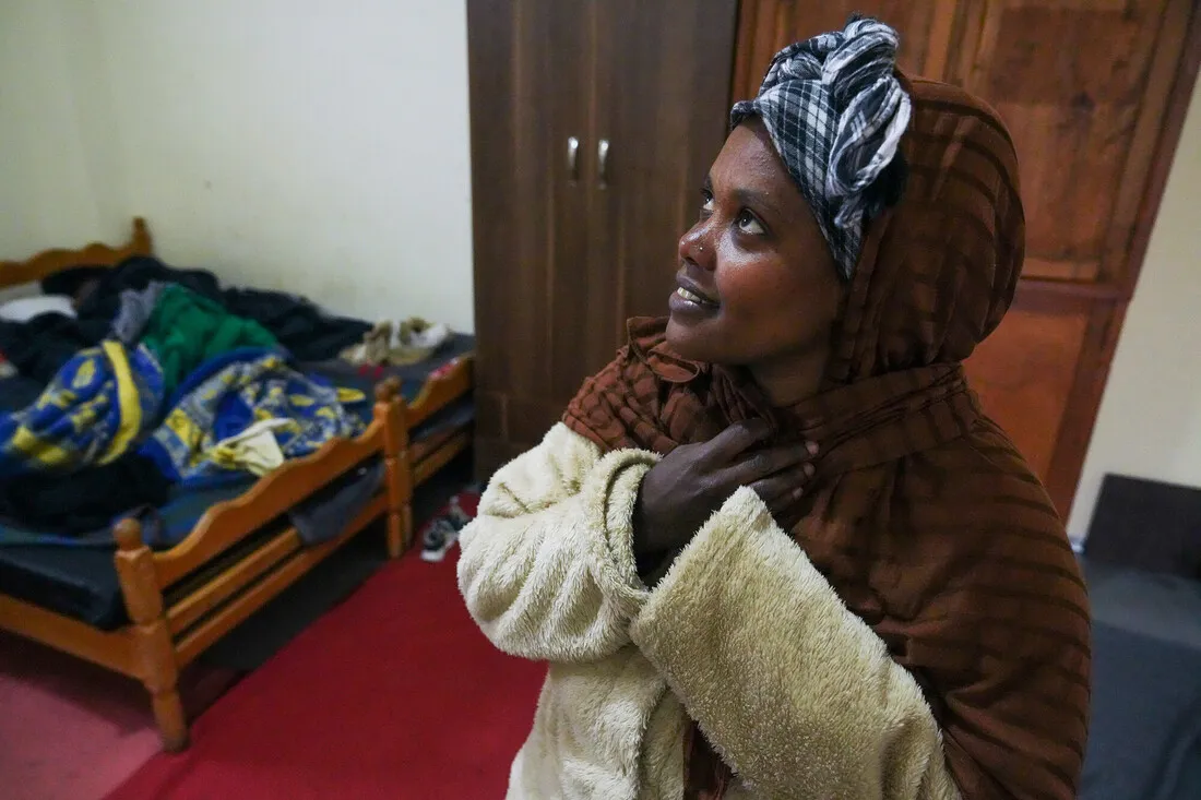 A woman wearing a hooded jacket and headscarf looks upward with a hopeful expression. She stands in a cozy room with a bed and wooden cupboard.