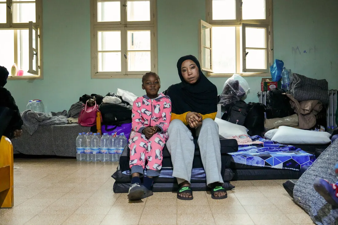 A woman in a black headscarf and yellow sweater sits on a mat beside a child in pink pajamas. Behind them are bags and water bottles. The room appears modest and lived-in, conveying a sense of resilience and togetherness.