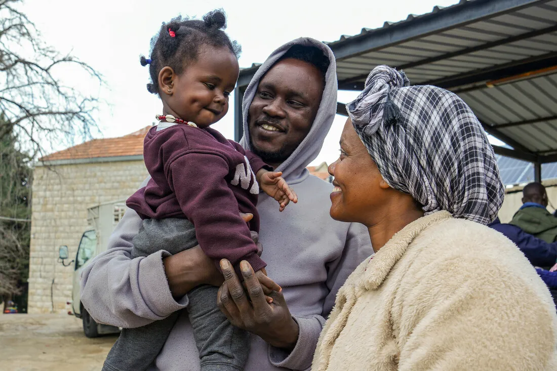 A smiling man holds a baby while a woman gazes at them warmly. They stand outdoors near a building, conveying a sense of family joy and togetherness.