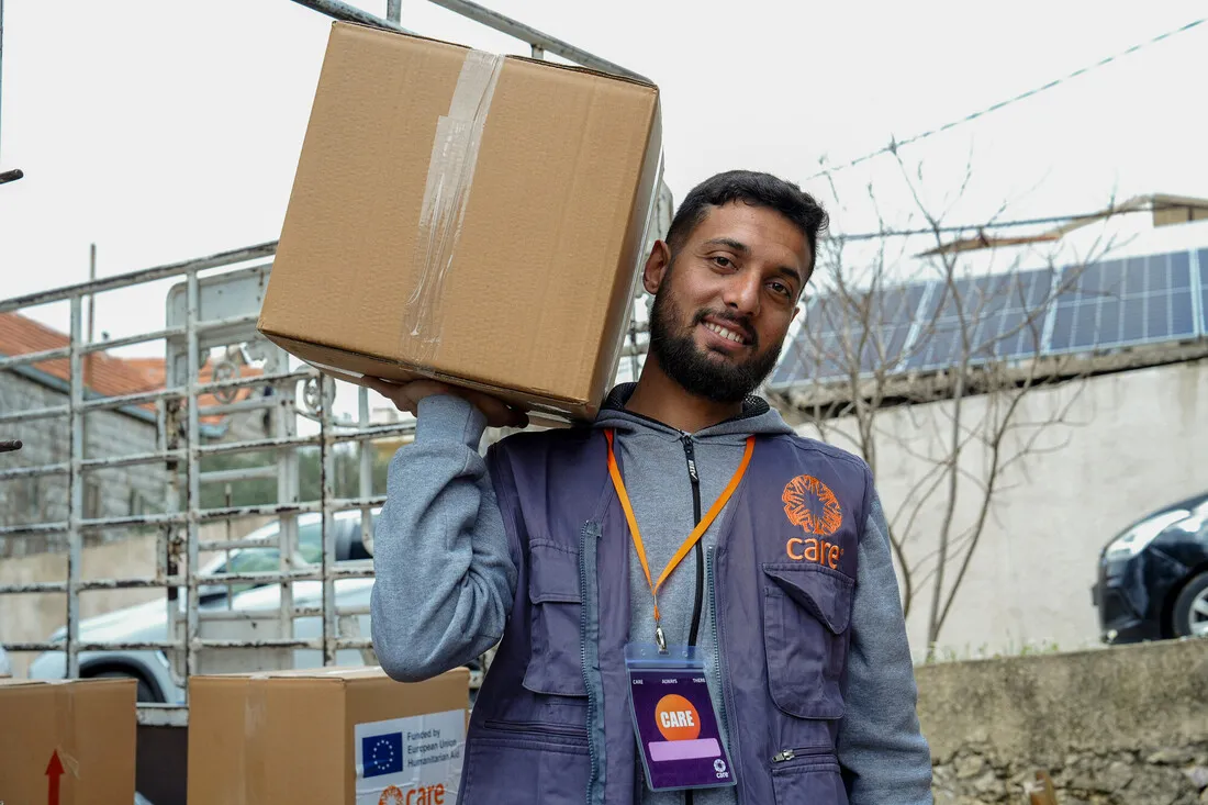 A man with a beard, wearing a purple vest and ID badge, smiles while carrying a cardboard box on his shoulder. Solar panels are visible in the background.