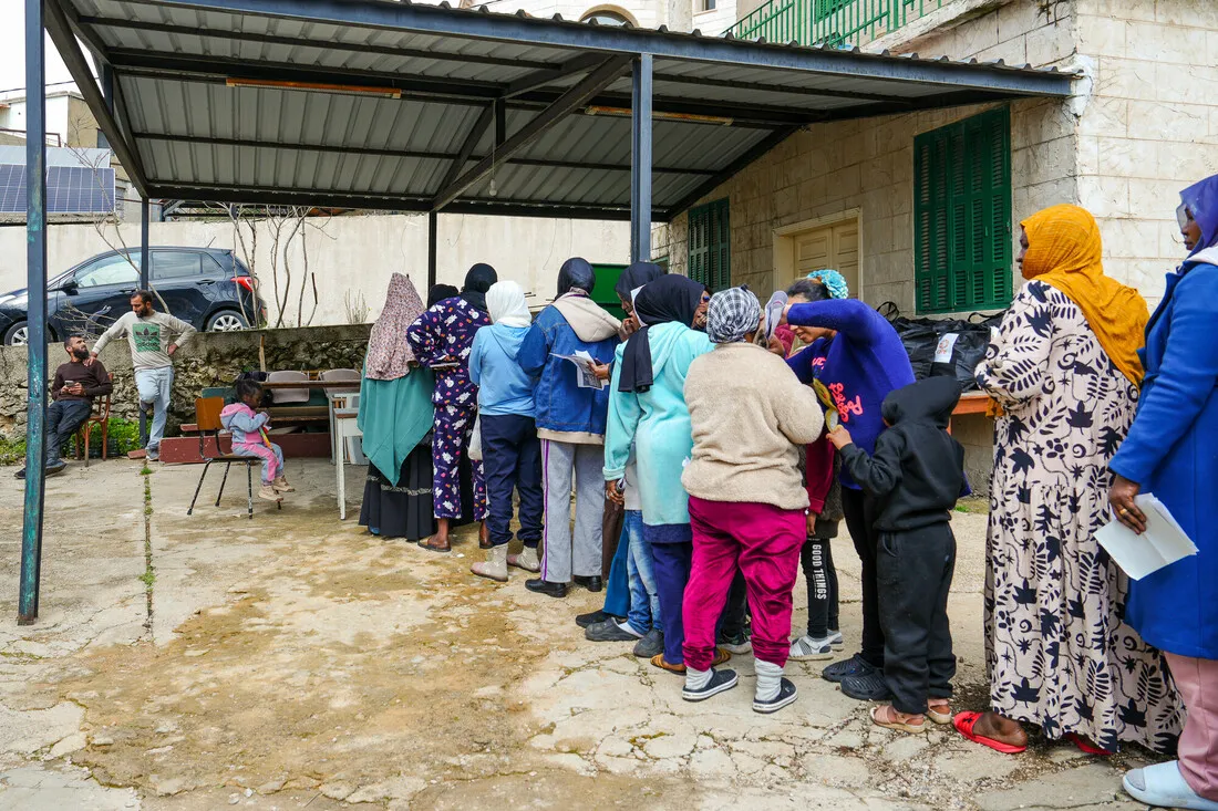 A diverse group of people wearing colorful clothing line up outdoors under a metal shelter. The tone is busy and communal, suggesting assistance or distribution.