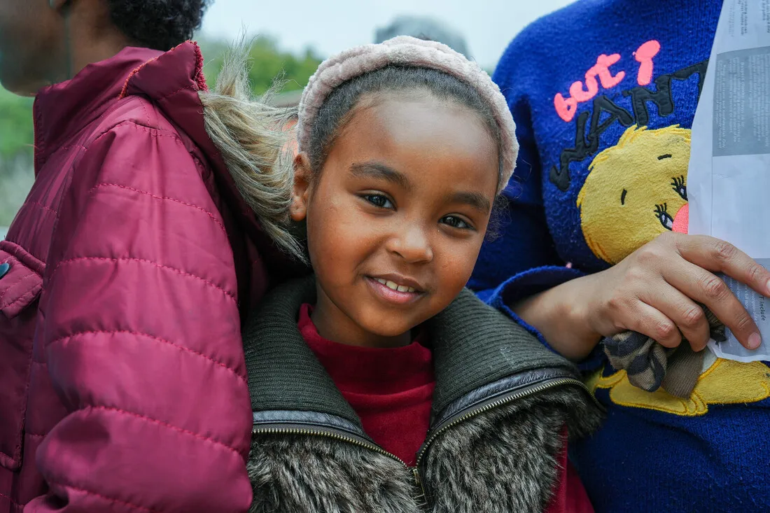 Young girl smiles warmly, wearing a fluffy headband and vest, standing between two people. One holds a paper, adding a cozy, cheerful vibe.
