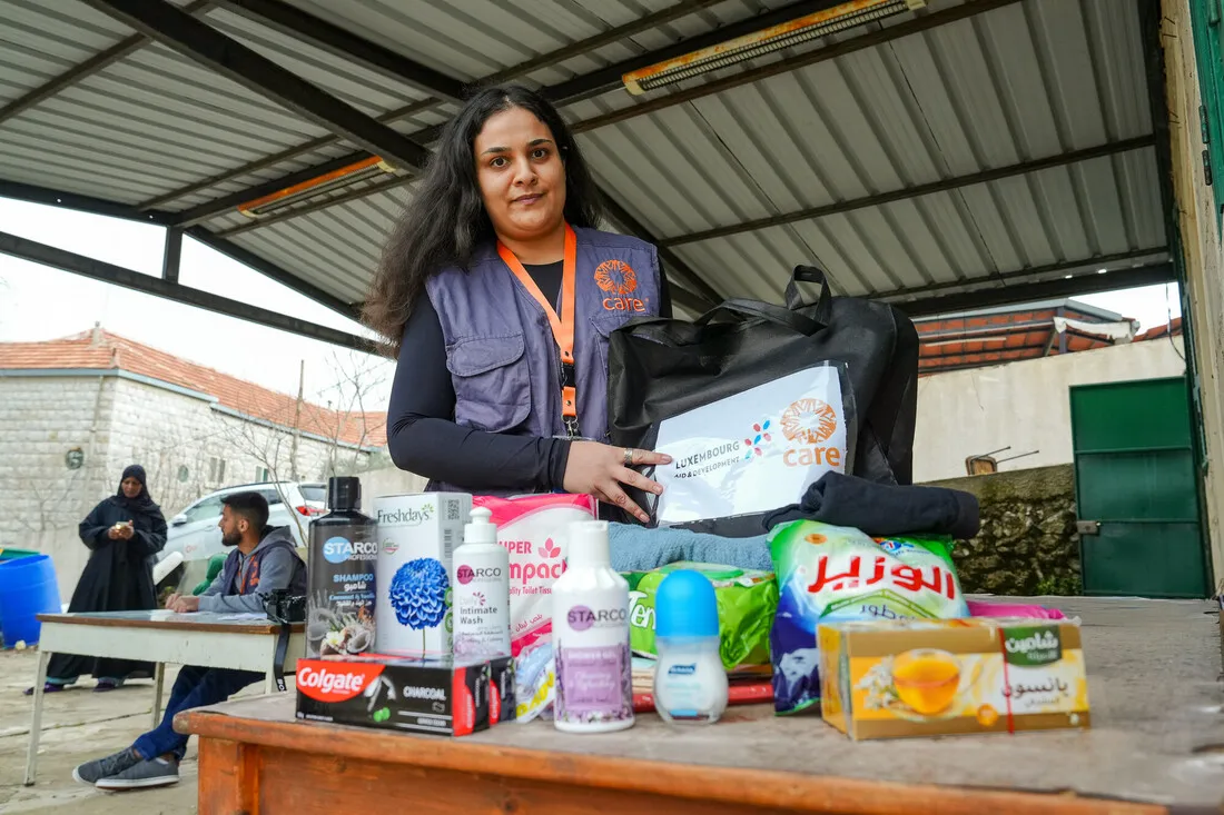 A woman wearing a CARE vest holds a black bag at a distribution point. Various personal care items are displayed on the table. The background has a few people and a metal roof, conveying a supportive and helpful atmosphere.