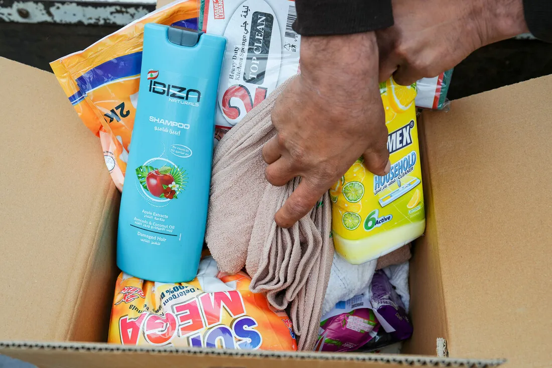 A cardboard box filled with household items including blue shampoo, yellow cleaning liquid, detergent, and a towel. Hands are organizing the contents.