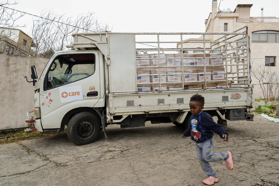 A child in motion passes a parked delivery truck loaded with CARE aid boxes. The scene appears to be in a neighborhood, evoking a sense of relief effort.