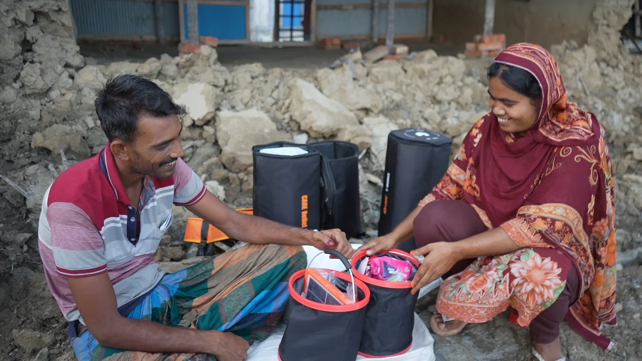 A man and a woman sitting amidst rubble, smiling as they look through the contents of two open CARE PACKAGE for Emergencies kits.