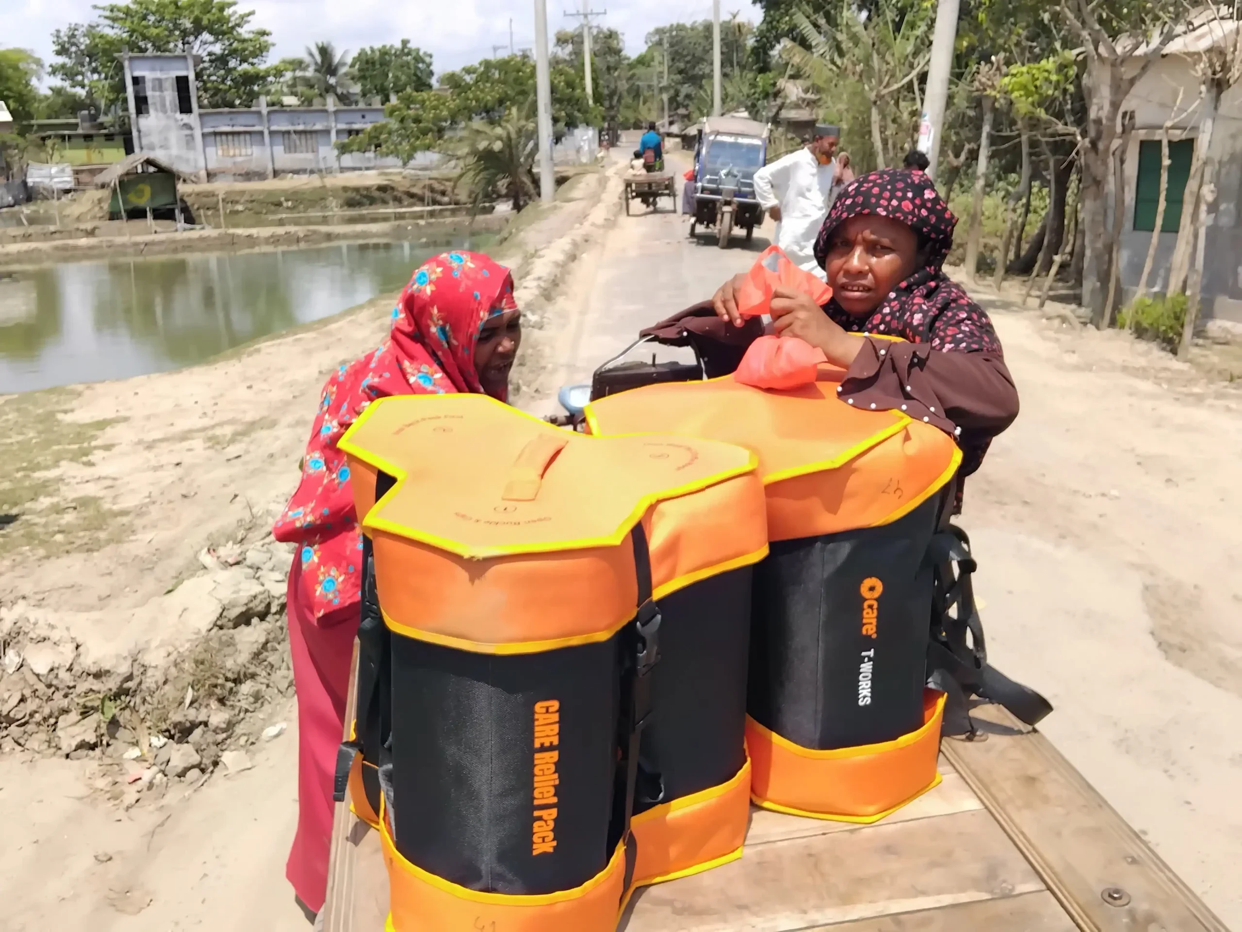 Two village women – CARE program participants – transport orange and black CARE Package for Emergencies kits on a three-wheeler rickshaw van along a rural road.